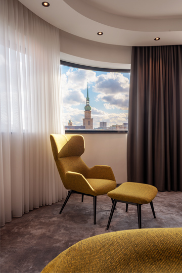Hotel room with a mustard-yellow armchair and footstool by a panoramic window, offering a view over Dortmund’s skyline.