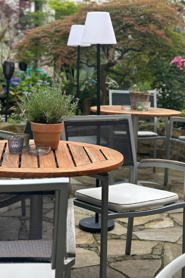 Cosy garden terrace with wooden tables, chairs, and potted plants surrounded by greenery.