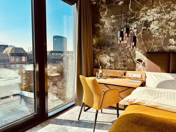 A bright hotel room with floor-to-ceiling windows, a view of the city, a bed, a desk, a yellow armchair and floral wallpaper.