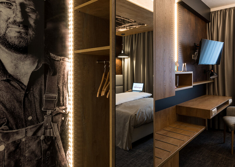 A modern hotel room with dark wood furnishings, an open-plan wardrobe, a mirror, a bed, a wall-mounted TV and warm LED strip lighting.