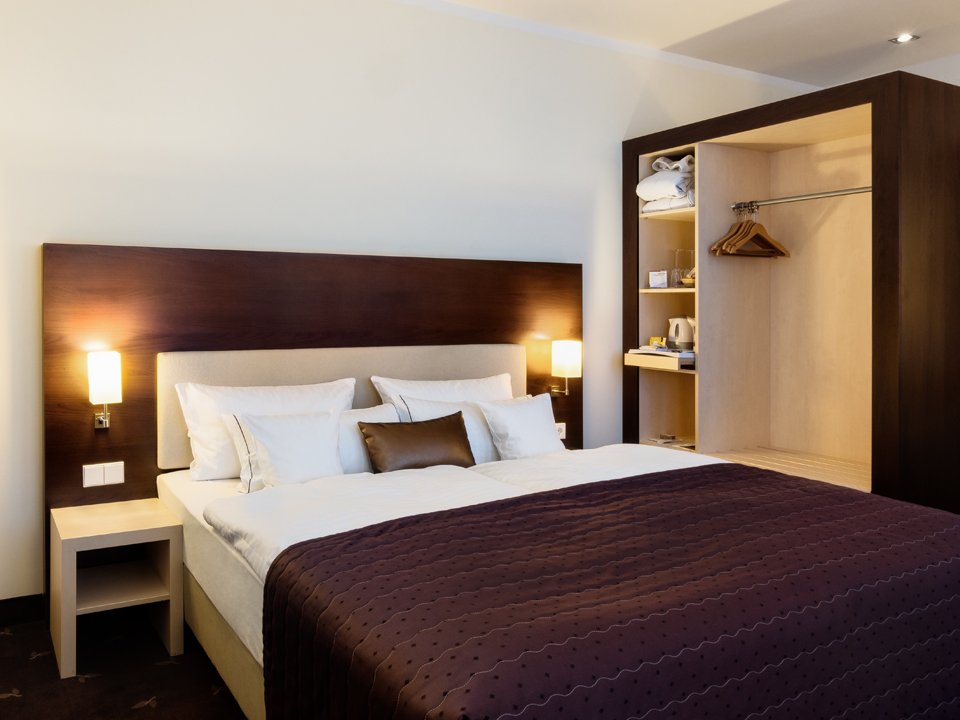 Hotel room with a double bed, bedside lamps, a dark headboard and an open wardrobe containing hangers, towels and a tea-making set.