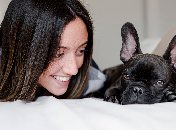 A smiling woman is lying on a bed, looking affectionately at a black French Bulldog lying next to her.