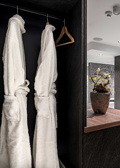 Two white bathrobes hanging in a dark wardrobe; next to them, a mirror reflecting a stylish vanity and a decorative plant.
