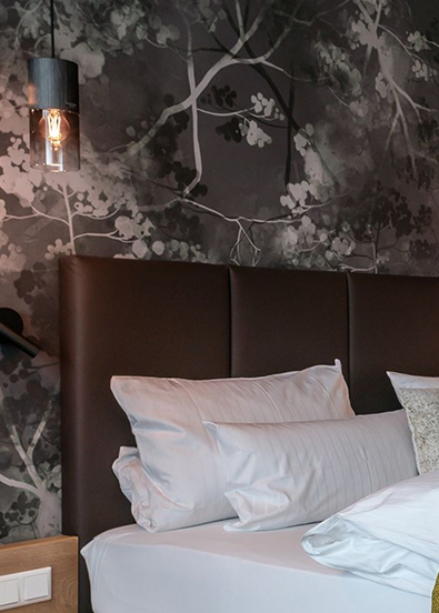 A bed in a modern hotel room with a brown headboard, white cushions, floral wallpaper and a hanging bedside lamp.