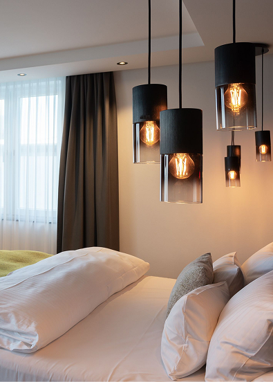A modern hotel room with a freshly made bed, large pillows, a window with curtains and decorative pendant lights giving off a warm glow.