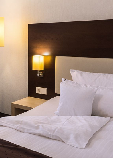 A modern hotel room with a freshly made bed, large pillows, a window with curtains and decorative pendant lights giving off a warm glow.