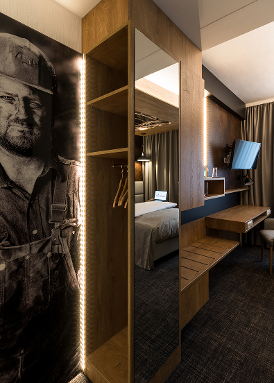 A modern hotel room featuring an open-plan wooden wardrobe, a full-length mirror, a bed, a desk and a large black-and-white wall mural depicting a worker.